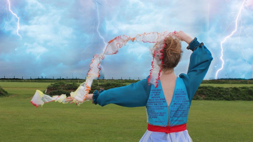 Model wearing student garments with their back to the camera. They are wearing a blue top and waving a fabric in the wind.