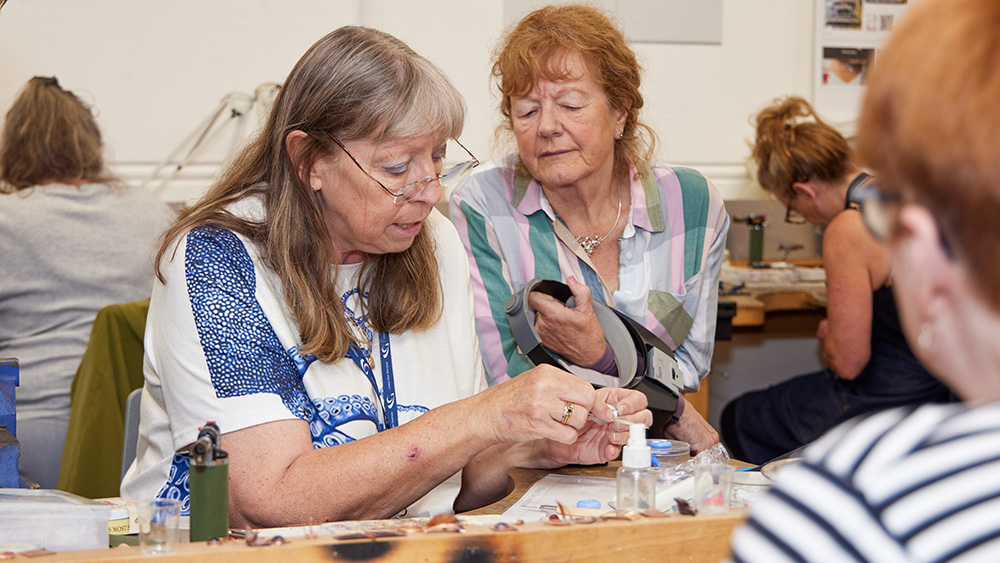 Adult learners watching silversmithing teacher demonstrate technique