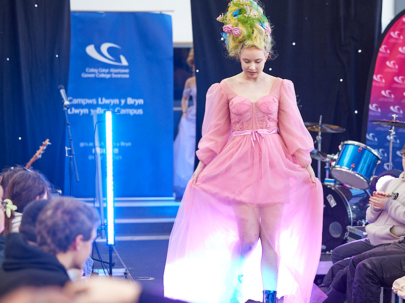 Model walking down catwalk wearing pink flowing dress and with tall pouf hair decorated with flowers