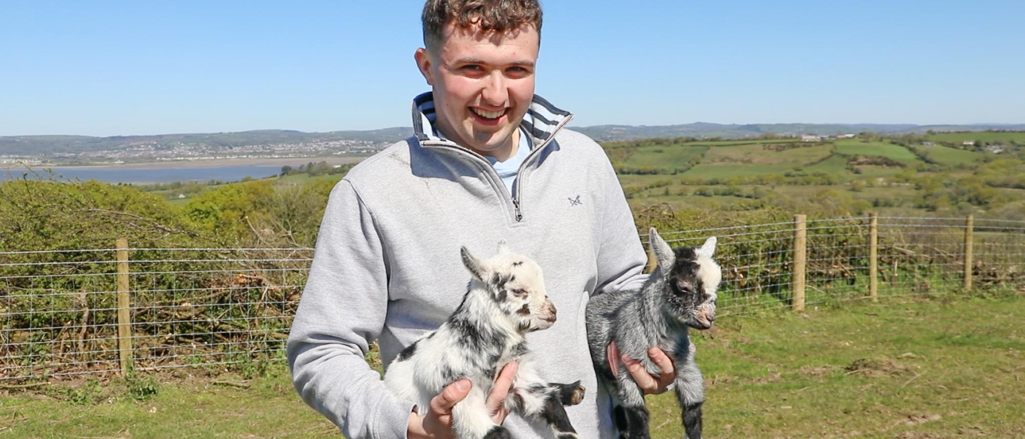 Will Evans yn dal dwy afr fach gyda golygfeydd Bro Gŵyr yn y cefndir