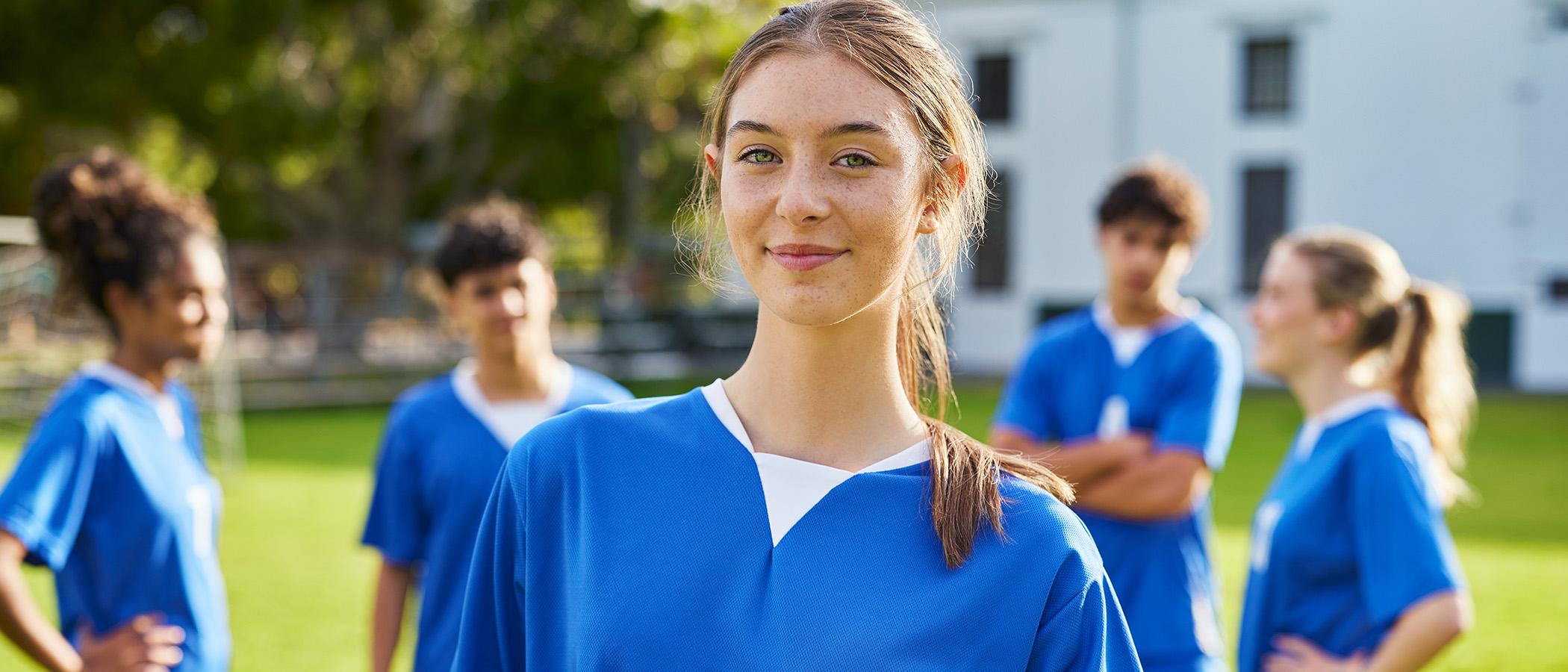 Teenager holds a football as she looks at the camera, with a group of other players behind her
