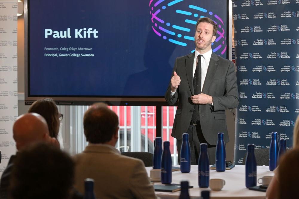 Man in grey suit, standing in front of presentation screen, talking