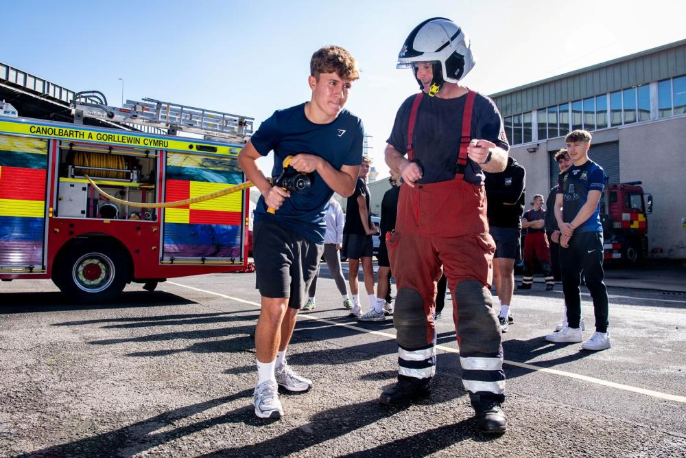 Student holding fire hose, with firefighter alongside, fire engine in background