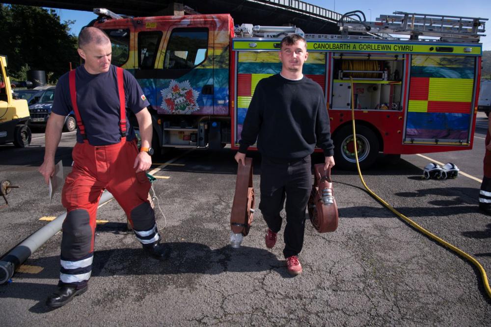 Student holding fire hose, with firefighter alongside, fire engine in background